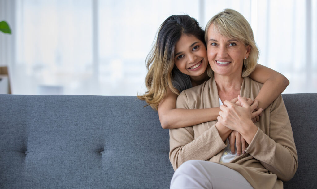 Delighted young Asian girl hugging happy middle aged foster mother sitting on sofa while spending time together at home
