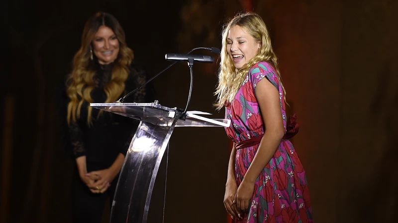 Child smiling and speaking at a podium, with an adult woman in the background, during an event supporting the Center for the Rights of Abused Children.