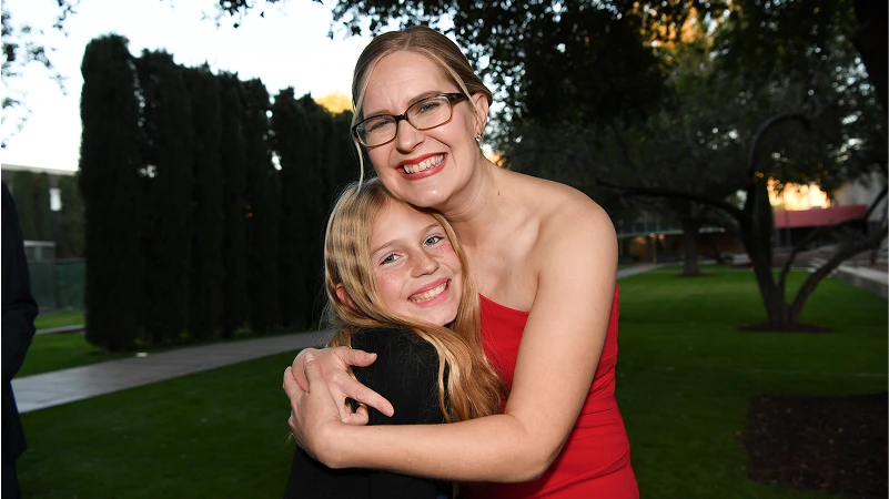 Nia Vardalos embracing a young girl, both smiling warmly, in an outdoor setting, highlighting a moment of joy and connection at a fundraising event for the Center for the Rights of Abused Children.