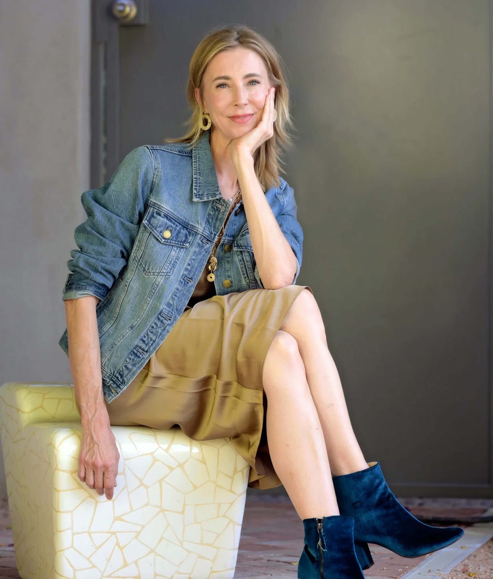 Woman in denim jacket and beige skirt sitting on a textured white stool, smiling and resting her chin on her hand, representing the Center for the Rights of Abused Children's advocacy efforts.