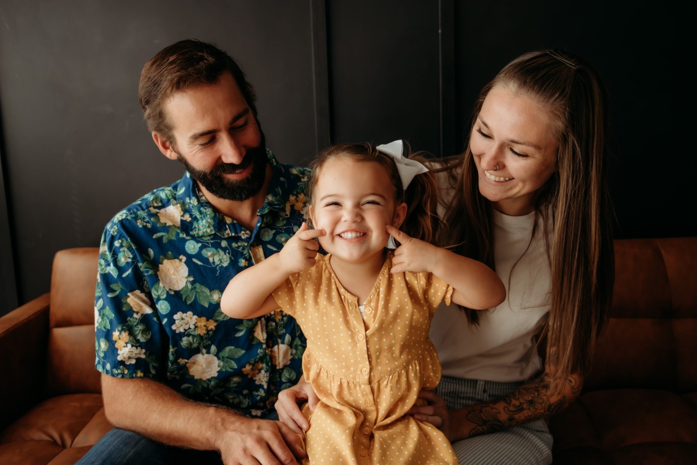 Smiling child making a silly face while sitting on a couch with two adults, representing family unity and joy, relevant to advocacy for permanency in foster care.