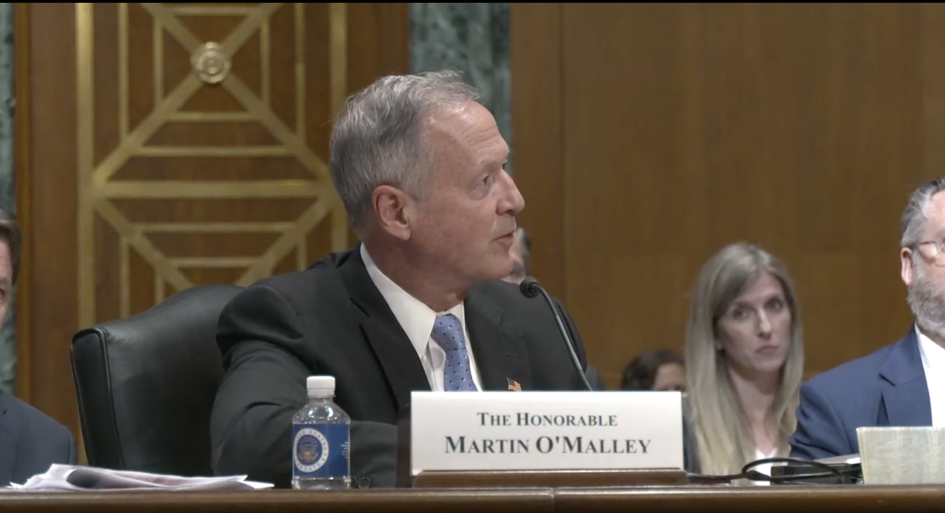 Honorable Martin O'Malley speaking at a Senate hearing on child welfare and foster youth benefits, with audience members in the background.